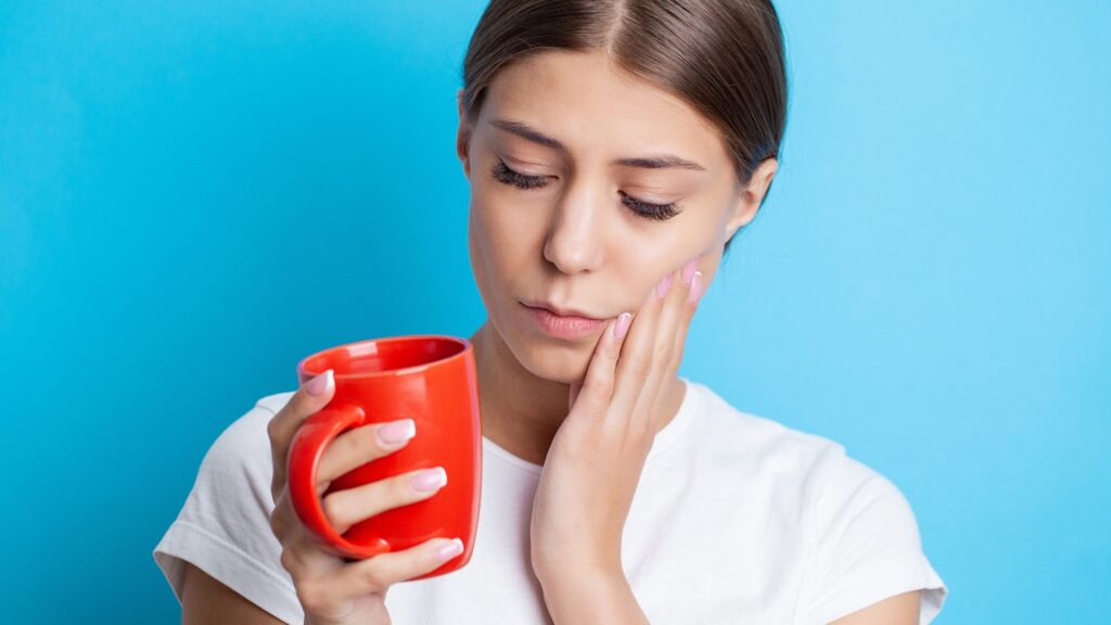 A girl is holding her mouth and a cup of tea, suffering from pain due to sensitive teeth