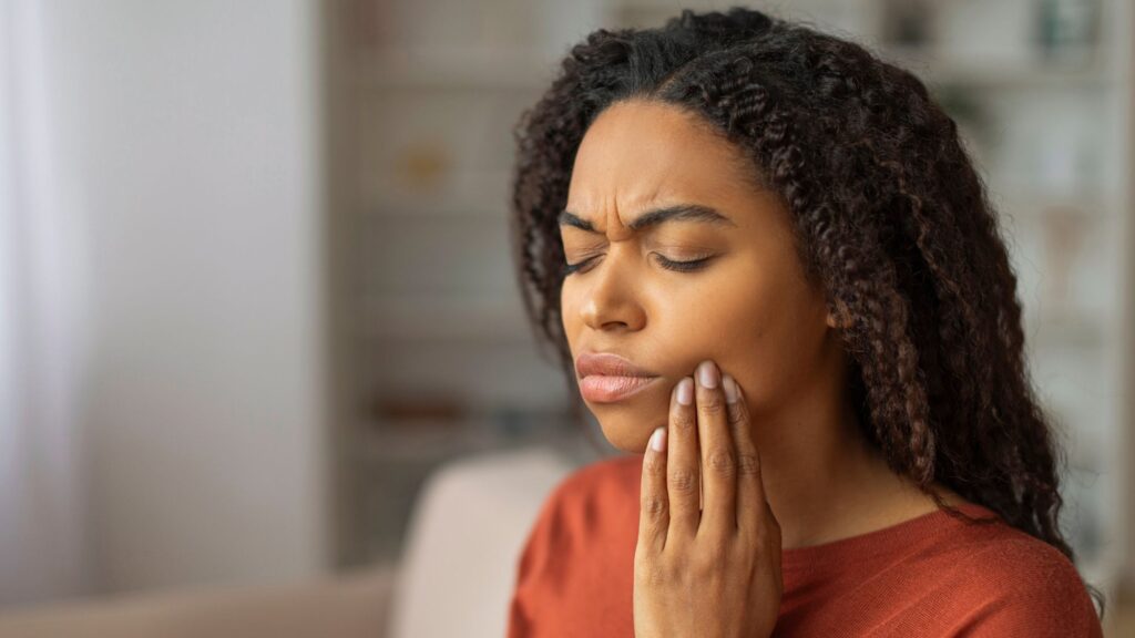 A young black woman holding her hand on her mouth, suffering from pain due to teeth sensitivity